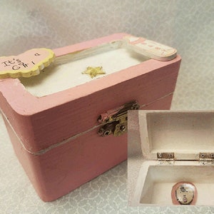 May include: A pink wooden box with a white interior and a small gold star on the lid. The box has a white string tied around it and a small heart-shaped tag that says "It's a Girl".