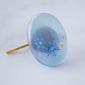 Può includere: Un accessorio per capelli azzurro con un perno color oro. L'ornamento ovale presenta disegni di fiocchi di neve blu e scaglie d'oro. L'accessorio è probabilmente realizzato in resina o materiale simile.