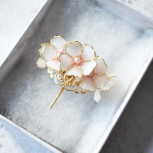 May include: A floral hair accessory featuring white flower petals with gold outlines and pink pearl accents. The hair piece has a gold stem and decorative filigree base, presented in a white box.