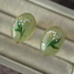 以下が含まれることがあります： 淡い緑色の楕円形イヤリングのペア。各イヤリングには、白い花、緑の葉、金色のアクセントで手描きされたスズランのデザインが施されています。イヤリングはニュートラルな背景に配置されています。