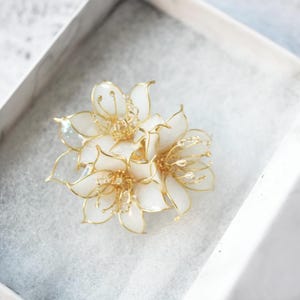 May include: A handcrafted floral brooch featuring white petals and gold-colored wire detailing. The flower is composed of multiple layers, creating a three-dimensional effect. The brooch is presented in a white box.
