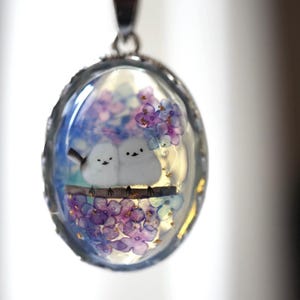 以下が含まれることがあります： 銀色のフレームの楕円形ペンダント。白い鳥のフィギュア2羽が枝に止まり、紫と青の花に囲まれています。ペンダントは透明な樹脂で満たされ、ミニチュアのシーンを作り出しています。