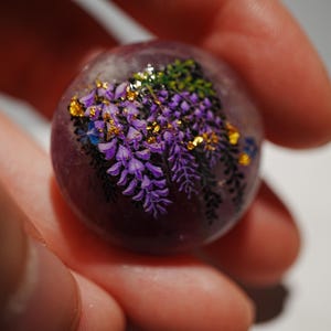 Può includere: Primo piano di una sfera di vetro viola con un motivo floreale. La sfera è decorata con fiori e foglie viola e dorati.