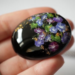Può includere: Una pietra decorativa nera di forma ovale con un motivo floreale. Presenta fiori di campanelle blu, viola e bianchi con foglie verdi e scaglie d'oro. La pietra ha una finitura liscia e lucida.
