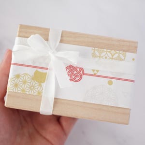 May include: A small, rectangular wooden box wrapped with a white ribbon and patterned paper. The paper features gold and red geometric designs. The box is tied with a white bow.