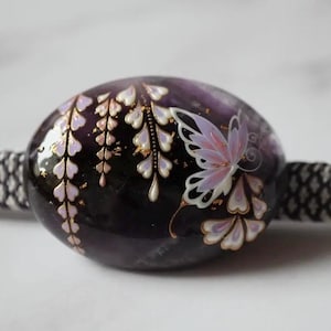 Può includere: Una perla ovale viola scuro con un disegno dipinto di farfalla e glicine. La farfalla ha ali rosa e bianche, e il glicine ha accenti bianchi e dorati. La perla è infilata su un cordoncino bianco e nero.