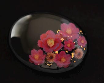 May include: A smooth, oval-shaped black resin piece featuring an embedded arrangement of pink flowers and small gold leaf flakes. The floral design is clustered on one side of the glossy, dark surface, showcasing various shades of pink petals.