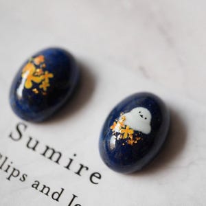 以下が含まれることがあります： 2つの濃い青色の楕円形のネイルアートデザイン。1つは金のフローラルデザイン、もう1つは金のフローラルデザインの白い雲が特徴です。「Sumire」という単語が背景に部分的に表示されています。