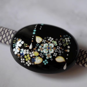 Può includere: Una perla decorativa nera di forma ovale con un motivo floreale. I fiori sono bianchi, azzurri e gialli con dettagli dorati. La perla è attaccata a un cordino fantasia.