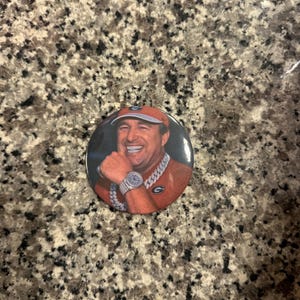 May include: A round button featuring a smiling man wearing a red baseball cap and a silver watch and chain. The man is wearing an orange shirt. The button is on a speckled brown surface.