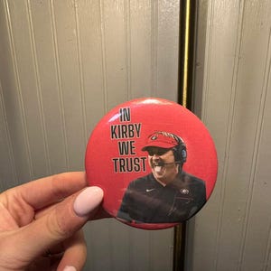 Puede incluir: Un botón rojo con el texto "IN KIRBY WE TRUST" y una imagen de un hombre con auriculares y una gorra roja. El botón es redondo y parece ser un pin o insignia.