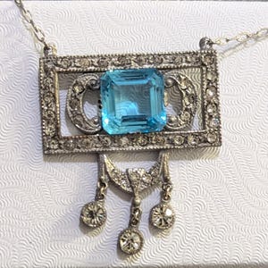 May include: A silver necklace with a pendant featuring a large blue gemstone surrounded by smaller clear gemstones. The pendant is in the shape of a rectangle with a decorative design.