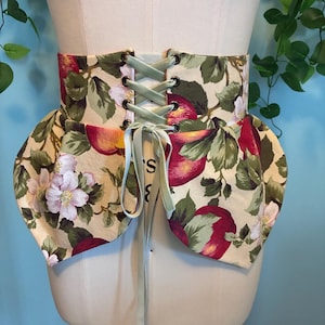 May include: A wide corset belt featuring a cream-colored fabric with a repeating pattern of red apples, green leaves, and pale pink blossoms. The belt includes bronze-toned metal eyelets and a light green satin ribbon for front lacing.