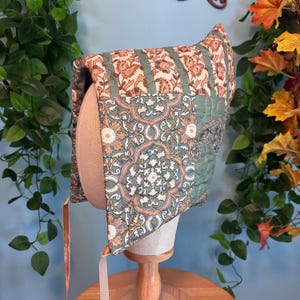 May include: A quilted bonnet featuring a patchwork design with sage green, cream, and burnt orange floral and geometric patterns. The headwear includes thin, light-colored fabric ties hanging from the bottom edges for securing under the chin.