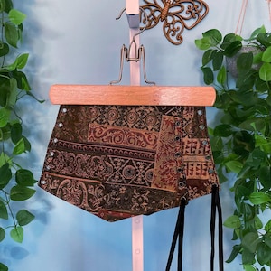 May include: A brown and green tapestry corset with black laces and eyelets. The corset is hanging on a wooden hanger.