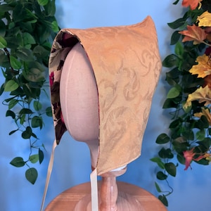May include: A bonnet featuring a gold-toned exterior with a subtle swirling jacquard pattern and a contrasting interior lining decorated with a dark floral print. The headwear includes thin, light-colored fabric ties attached at the base.