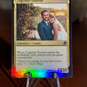 K&ouml;nnte beinhalten: Eine Regenbogen-Foil-Sammelkarte mit dem Titel Together Forever und einem Foto eines Paares in Hochzeitskleidung. Der Kartentext enth&auml;lt Legendary - Couple, Spielregeln und ein Zitat von Victor Hugo. Unten rechts sind Unendlichkeitssymbole zu sehen.
