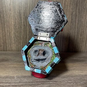 May include: A hexagonal wooden box with a black and white checkered pattern on the outside. The box is open and reveals a small, gray, sculpted figure of a creature with large eyes and a wide mouth. The figure is surrounded by green glitter.
