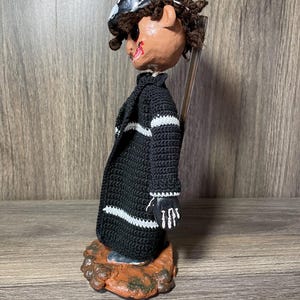 May include: A handmade doll with a black and white striped sweater, black hat, and a painted face. The doll is standing on a brown base.