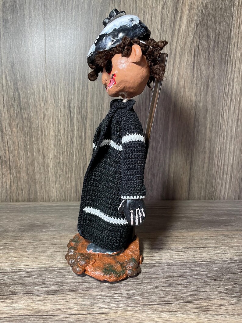 May include: A handmade doll with a black and white striped sweater, black hat, and a painted face. The doll is standing on a brown base.