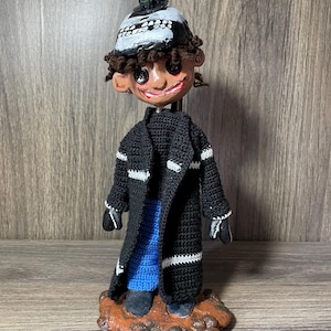 May include: A handmade doll with a black skull hat, black and white striped coat, blue pants, and brown shoes. The doll is standing on a brown base.