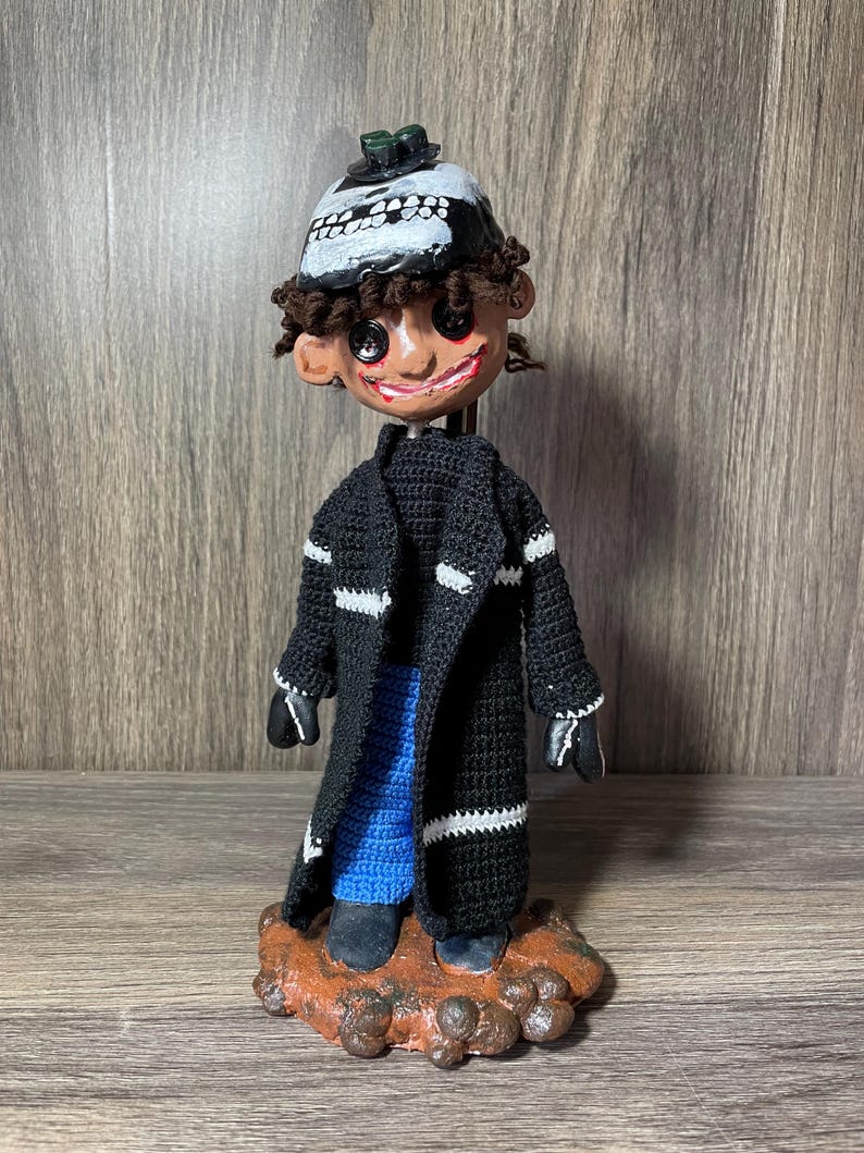 May include: A handmade doll with a black skull hat, black and white striped coat, blue pants, and brown shoes. The doll is standing on a brown base.