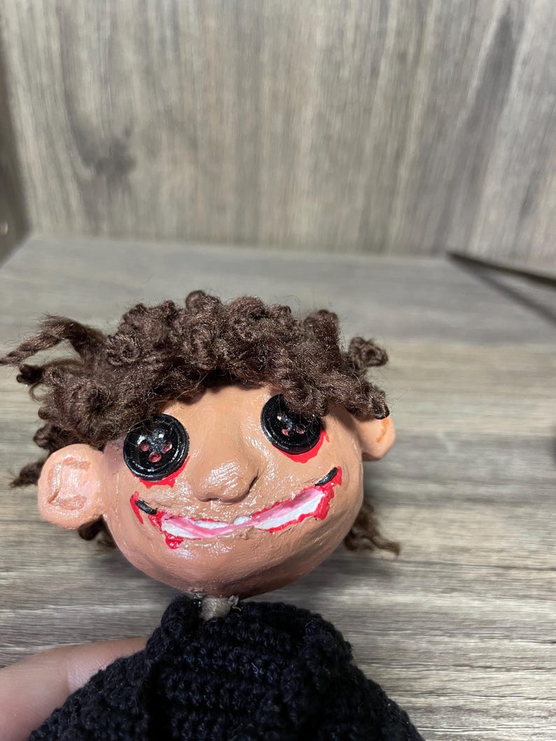 May include: Handmade doll with brown yarn hair, a painted face with red and black details, and a black knitted sweater.