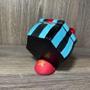 May include: A small, hexagonal wooden box with a blue and black striped design. The box has a red ribbon and a silver hinge. The box is sitting on a red, star-shaped object.