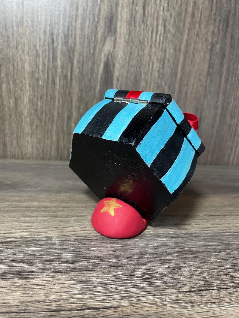 May include: A small, hexagonal wooden box with a blue and black striped design. The box has a red ribbon and a silver hinge. The box is sitting on a red, star-shaped object.