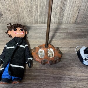 May include: A handmade doll with a black and white striped sweater, blue pants, and a mischievous smile. The doll is standing on a brown base with a metal rod. A white skull with green eyes is also pictured.
