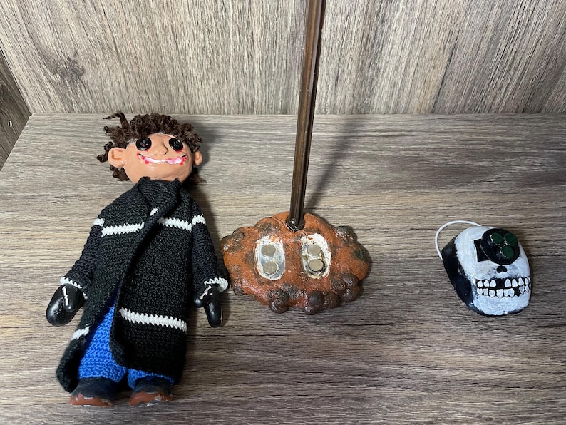 May include: A handmade doll with a black and white striped sweater, blue pants, and a mischievous smile. The doll is standing on a brown base with a metal rod. A white skull with green eyes is also pictured.
