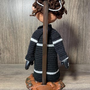 May include: A black and white crocheted doll with brown curly hair wearing a black and white hat. The doll is standing on a brown base with a wooden dowel.