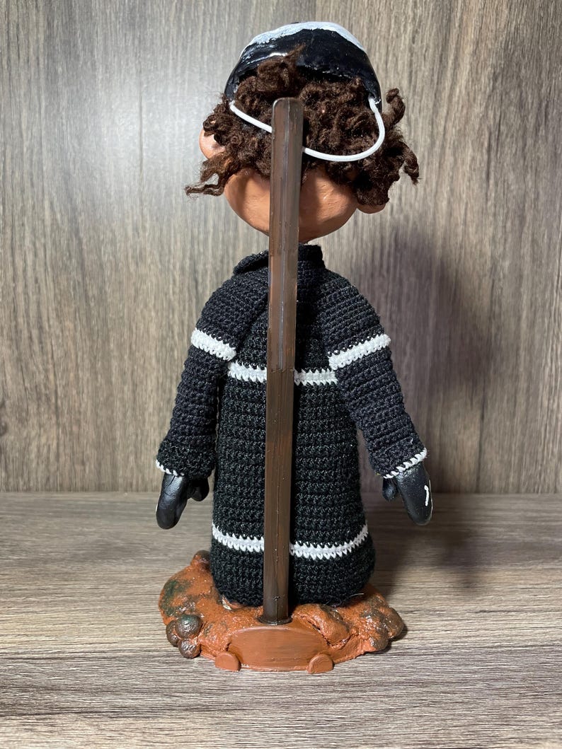 May include: A black and white crocheted doll with brown curly hair wearing a black and white hat. The doll is standing on a brown base with a wooden dowel.