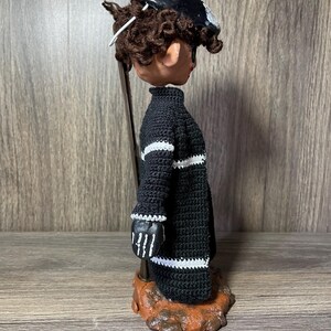 May include: A black and white crocheted doll with a black and white hat. The doll is standing on a brown base.