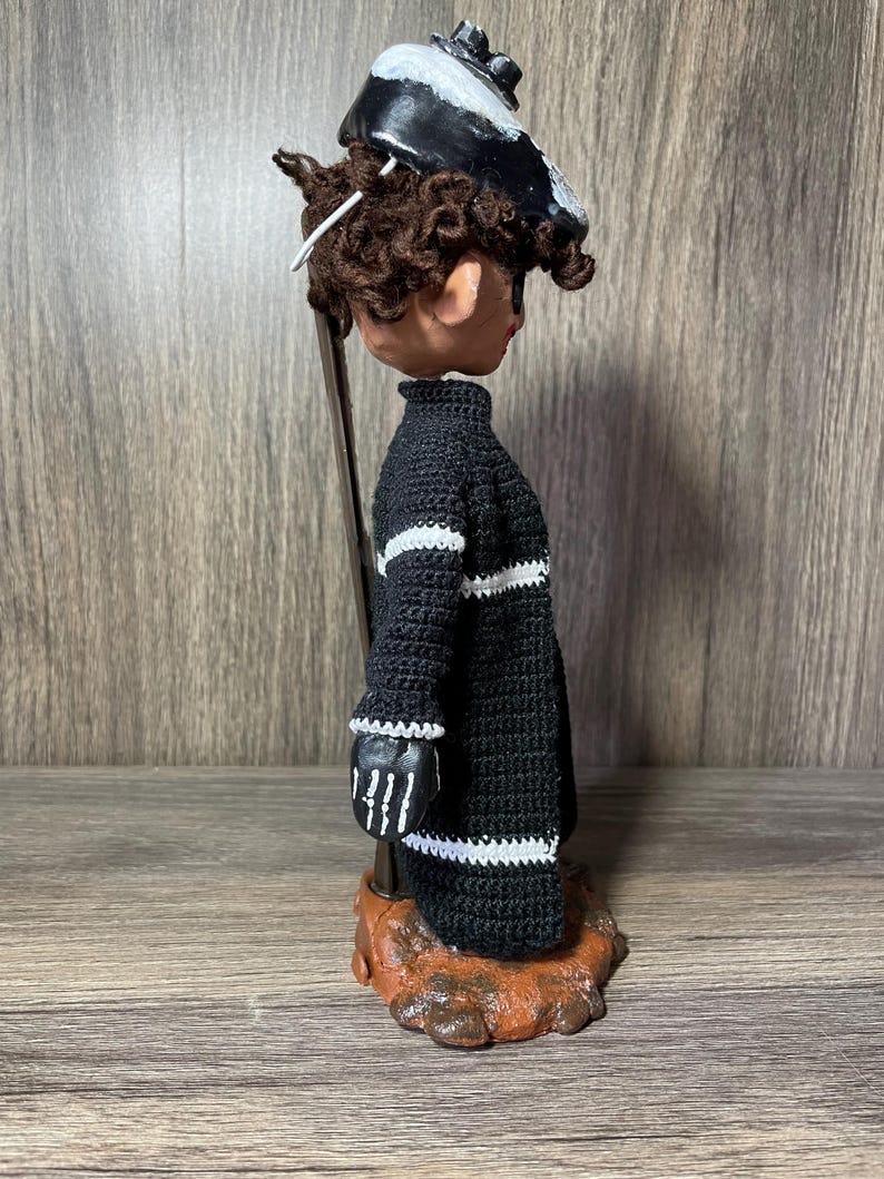 May include: A black and white crocheted doll with a black and white hat. The doll is standing on a brown base.