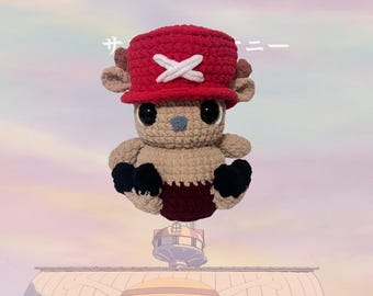 May include: A crocheted plush toy reindeer wearing a red hat with a white X on it. The reindeer is brown and has black eyes and a black nose. The reindeer is sitting on a white background.