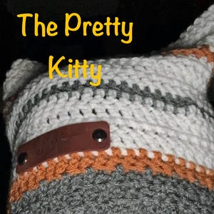 May include: A crocheted cat hat with a brown leather patch and metal poppers. The hat is striped with white, grey, and orange yarn.