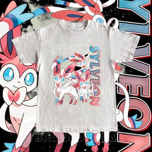 May include: A light gray t-shirt featuring a Sylveon graphic. The design includes multiple Sylveon characters in pink, white, and blue, with the word "Sylveon" in a gradient font. The background is black with white and pink accents.