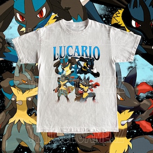 May include: A light gray t-shirt featuring the word "LUCARIO" in blue, with a graphic of multiple Lucario characters in various poses. The design includes blue, black, yellow, and red accents. The shirt has a crew neck and short sleeves.