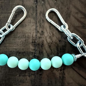 Może przedstawiać: Srebrny metalowy łańcuch z dwoma karabińczykami i rzędem turkusowych i jasnoniebieskich koralików. Koraliki są okrągłe i równomiernie rozmieszczone wzdłuż łańcucha, tworząc element dekoracyjny.