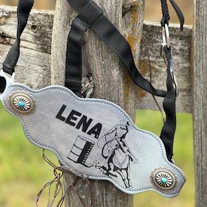 May include: A silver leather halter with a turquoise stone design and the name "LENA" engraved on it. The halter features a black and white illustration of a barrel racer on a horse.