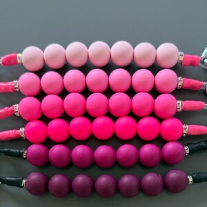 Puede incluir: Seis cordones con cuentas dispuestos en un degradado de rosa claro a morado oscuro. Cada cordón presenta siete cuentas redondas de silicona, espaciadores de metal plateado con cristales transparentes y un cordón de color sólido en los extremos.