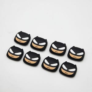 May include: Eight black Batman-themed shoe charms. Each charm features a black mask with white eyes and a tan mouth. The charms are arranged on a white surface.