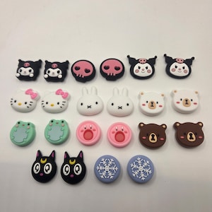 May include: A collection of decorative shoe charms in various designs. The charms include cartoon characters such as Hello Kitty, bunnies, bears, frogs, and cats. Other designs include skulls, pink circles, and snowflake patterns. The charms are in various colors, including black, white, pink, and blue.