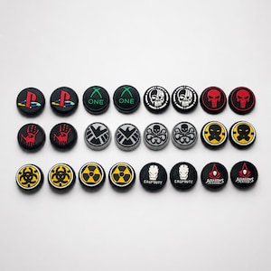 May include: A collection of black controller thumb grips with various designs. The grips feature symbols like the PlayStation logo, Xbox logo, skulls, and biohazard symbols. Some grips have the text "ONE" and "Call of Duty".