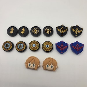 May include: A collection of controller thumb grips and decorative accessories. The set includes black and gold circular grips with symbols, shield-shaped grips in black and gold, and blue and red, plus two character-shaped grips.