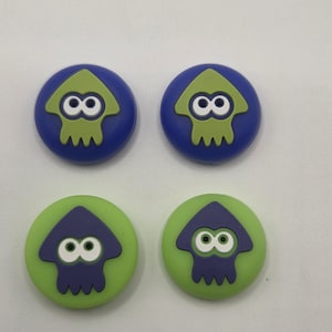 May include: Four circular buttons with a cartoon squid design. Two buttons have a blue background with a green squid, and two have a green background with a purple squid. The squid has white eyes.