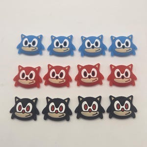 May include: An assortment of twelve cartoon character charms in three colors: blue, red, and black. Each charm features a smiling face with large eyes and a tan belly. The charms are arranged in three rows of four.