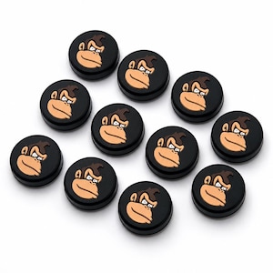 May include: A collection of black, circular buttons featuring a cartoon monkey face with a tan face and brown hair. The monkey has a stern expression. The buttons are arranged on a white background.