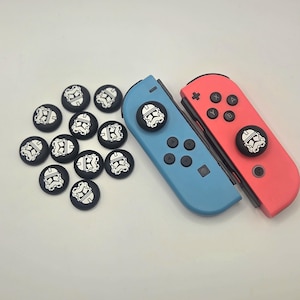 May include: A set of twelve black circular thumbstick covers featuring a white printed clone trooper helmet design, alongside one blue and one red game controller, with two of the covers attached to the controller joysticks.